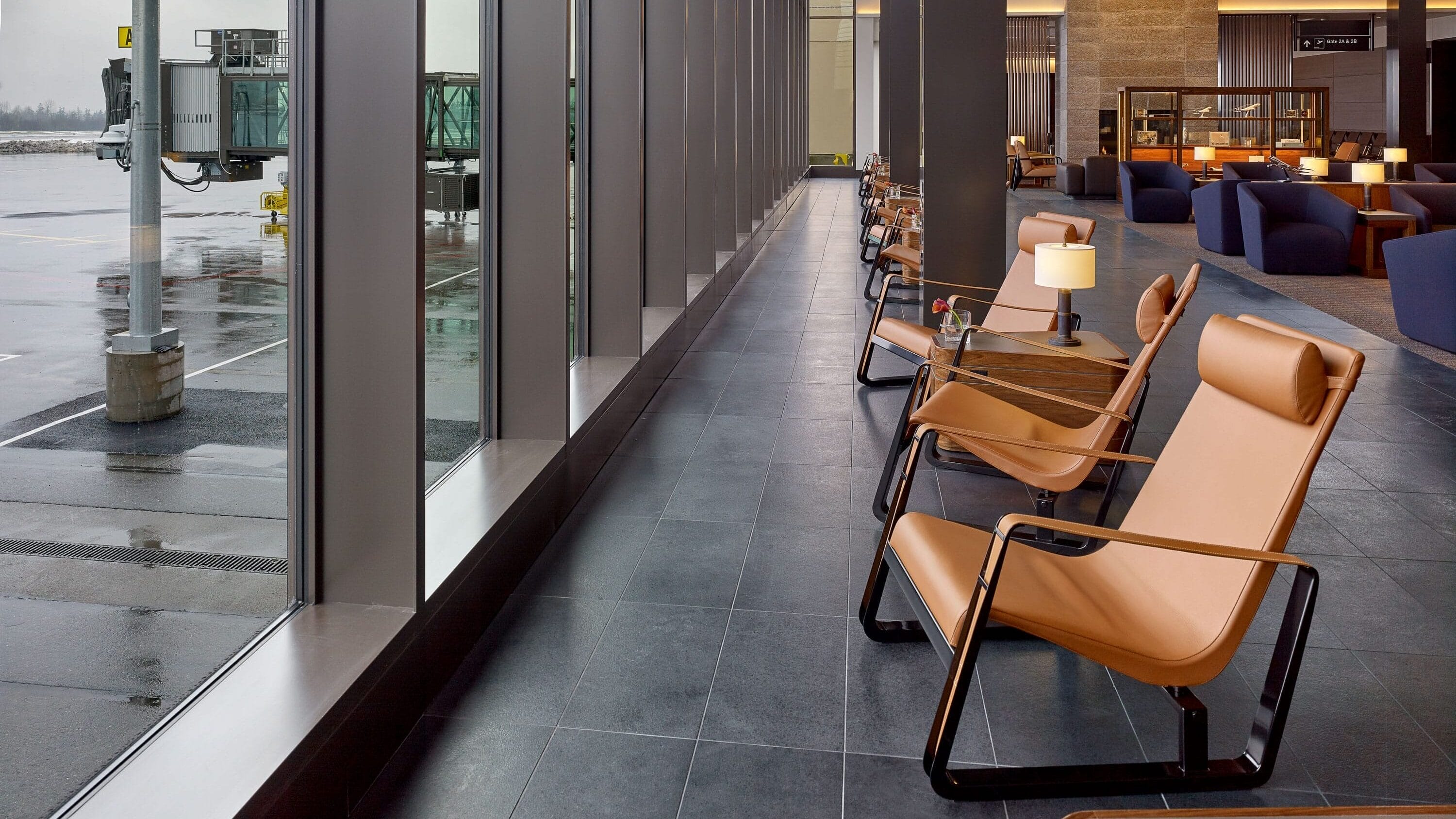 Interior of Paine Field passenger terminal lounge with floor-to-ceiling windows overlooking the airport apron, leather lounge chairs and tables, and a modern wood-paneled ceiling