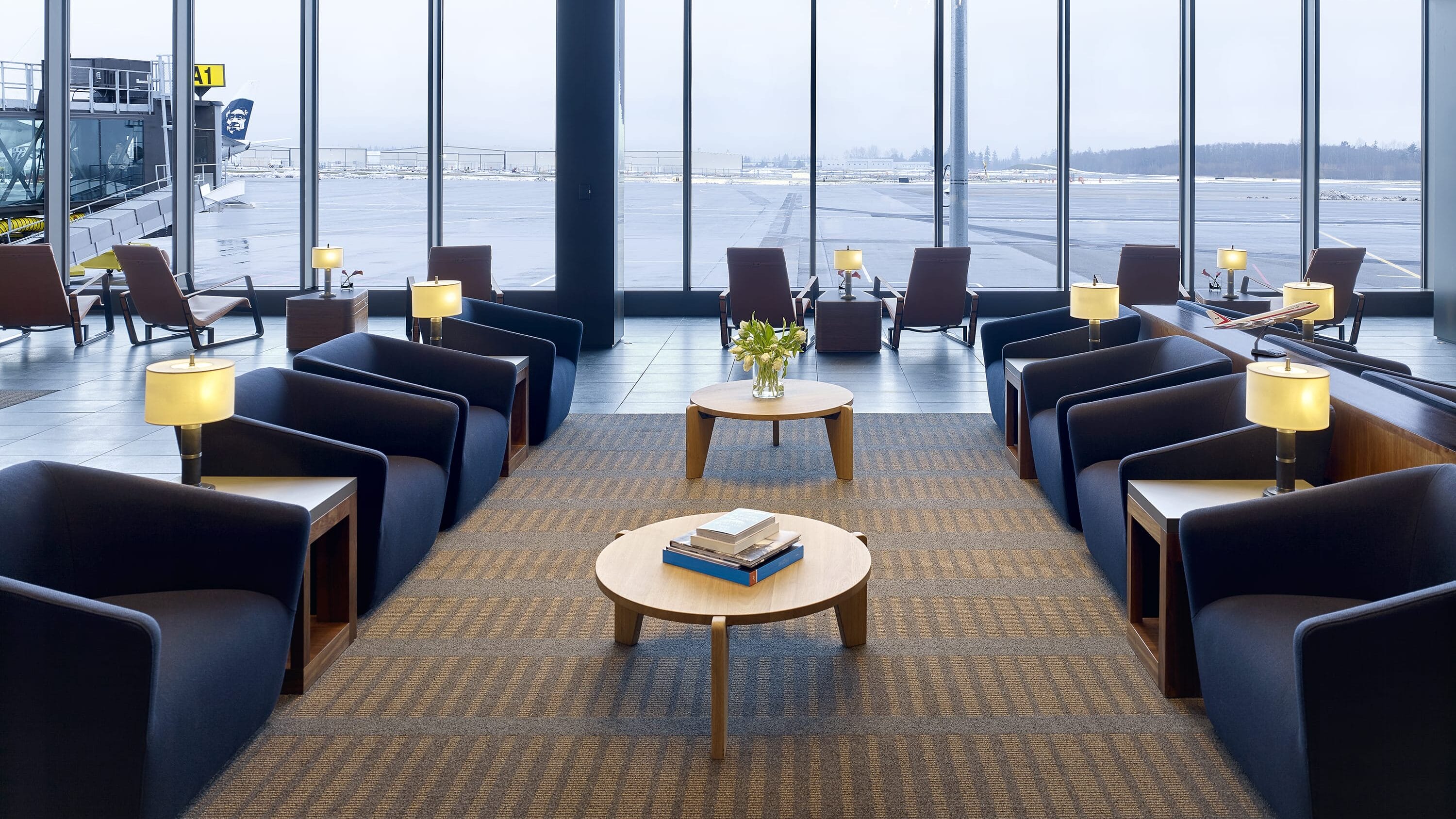 Interior of Paine Field passenger terminal lounge with floor-to-ceiling windows overlooking the airport apron, dark navy lounge chairs and wood accent tables